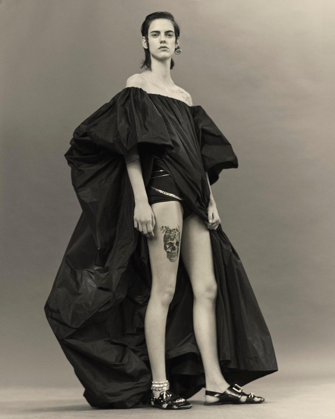 Fashion shot of a female model in a dramatic black gown with a skull tattoo on her thigh, posing against a neutral grey backdrop.