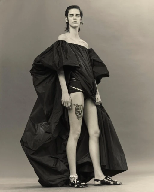 Fashion shot of a female model in a dramatic black gown with a skull tattoo on her thigh, posing against a neutral grey backdrop.