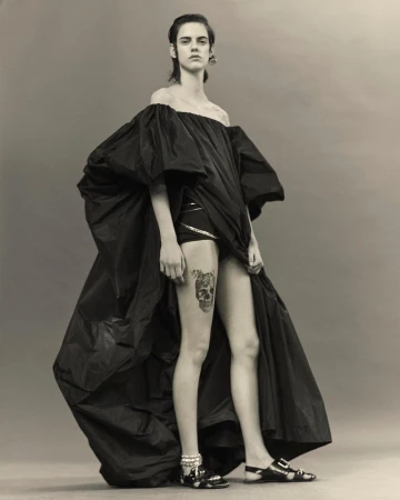 Fashion shot of a female model in a dramatic black gown with a skull tattoo on her thigh, posing against a neutral grey backdrop.
