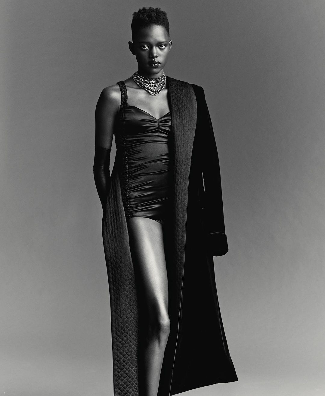 Monochrome portrait of a Black woman with short hair wearing a necklace, bodysuit, gloves and robe against a grey backdrop.