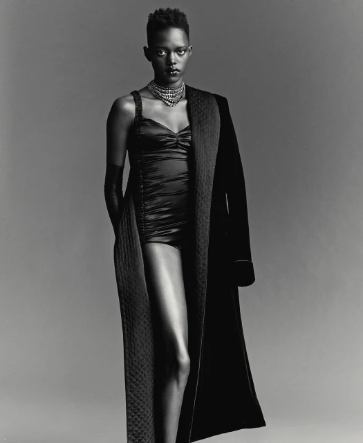 Monochrome portrait of a Black woman with short hair wearing a necklace, bodysuit, gloves and robe against a grey backdrop.