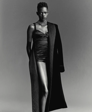 Monochrome portrait of a Black woman with short hair wearing a necklace, bodysuit, gloves and robe against a grey backdrop.