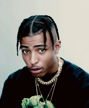 Close-up portrait of an adult male with braided hair wearing a Hulk graphic t-shirt and gold chains against a neutral backdrop.