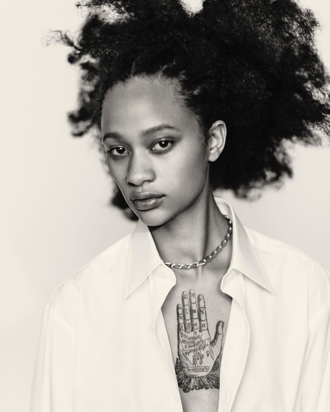 Striking black and white portrait of a young adult woman with an afro, wearing a button-down shirt and silver chain necklace against a white backdrop.