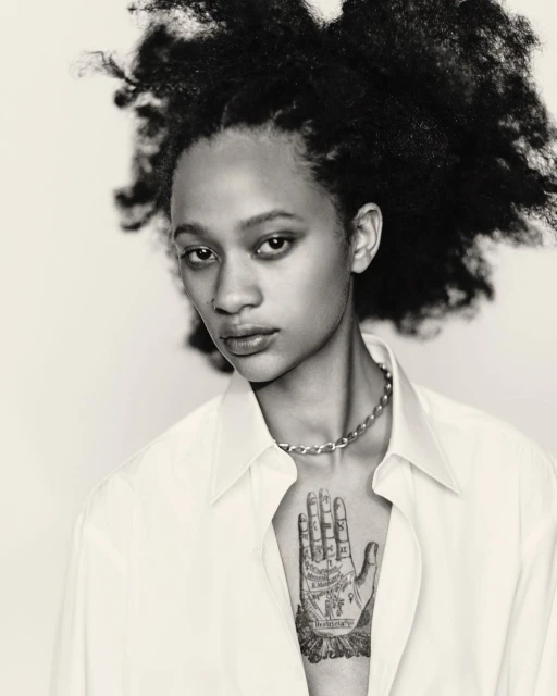 Striking black and white portrait of a young adult woman with an afro, wearing a button-down shirt and silver chain necklace against a white backdrop.