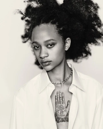 Striking black and white portrait of a young adult woman with an afro, wearing a button-down shirt and silver chain necklace against a white backdrop.