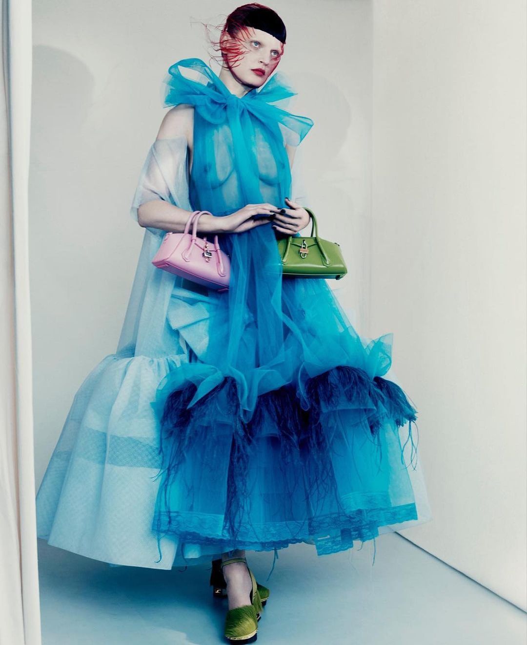 A woman in a large, layered blue tulle dress holds a pink purse in one hand and a green purse in the other against a light background.