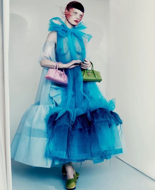 A woman in a large, layered blue tulle dress holds a pink purse in one hand and a green purse in the other against a light background.