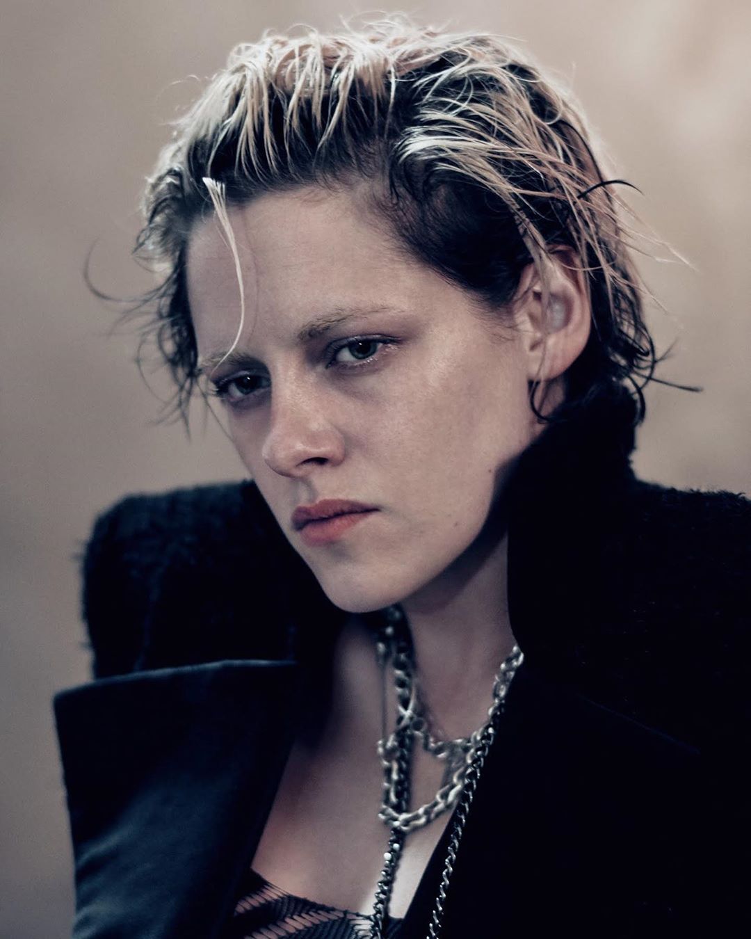 Portrait of an adult female with short, light-streaked hair and dark eye makeup, wearing a black top with a chain necklace in a studio.