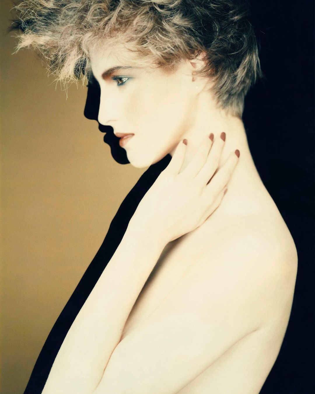 Profile portrait of a young fair-skinned woman with blonde curly hair, painted red nails, and bare shoulders against a two-tone background.