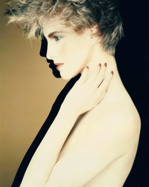Profile portrait of a young fair-skinned woman with blonde curly hair, painted red nails, and bare shoulders against a two-tone background.