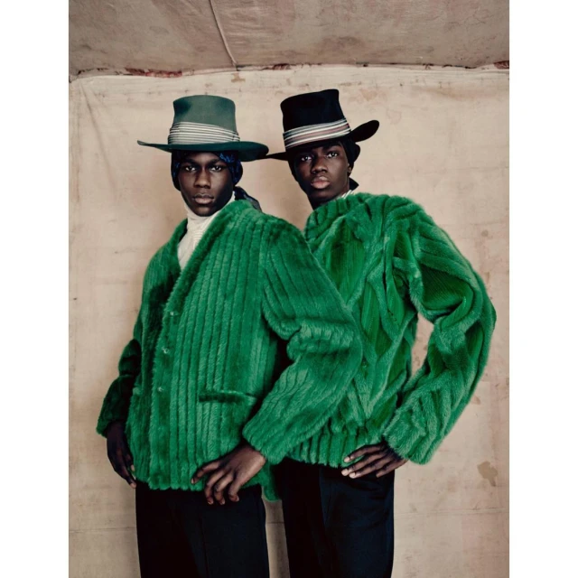 Two Black men in green fur jackets and hats pose indoors against a neutral backdrop, showcasing fashion-forward, textured outerwear.