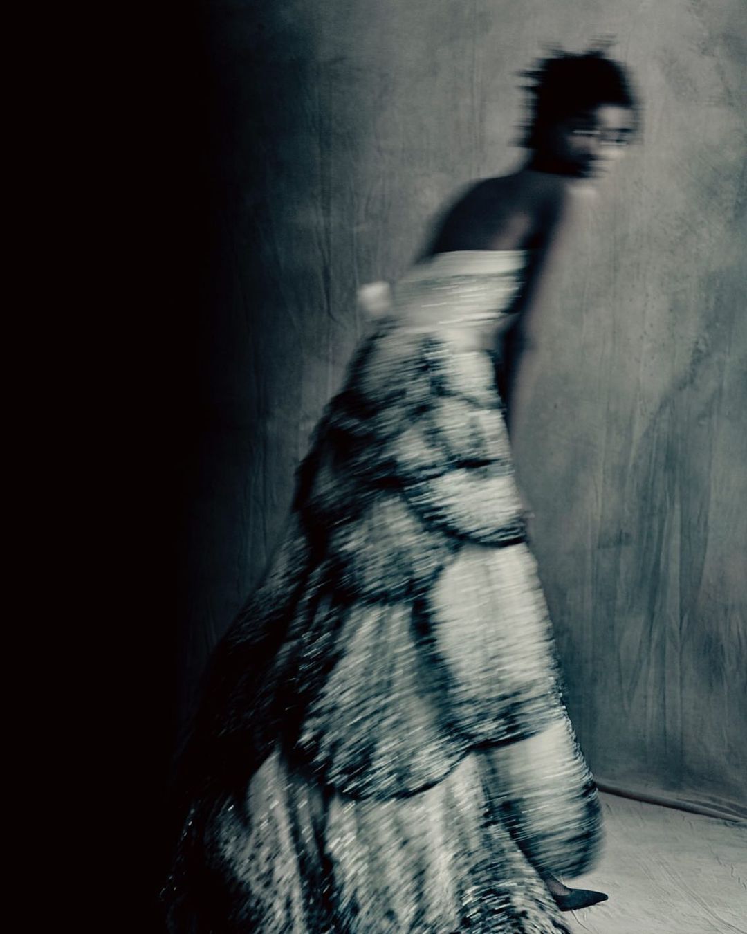 Woman in motion wearing a long, layered dress in a blurred photo studio setting with dark shadows and a neutral background.