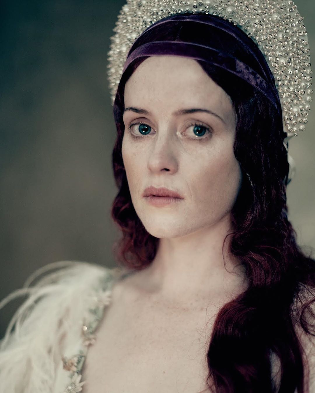 Portrait of a woman with red hair, wearing a jeweled headpiece and feathered top, set against a blurred neutral background.
