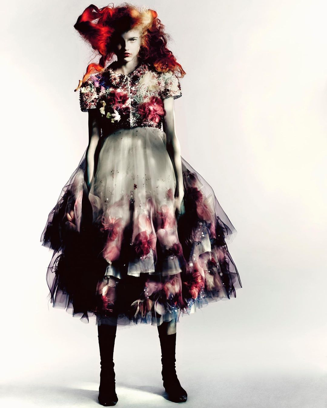 Fashion shot of a young female model in an ornate, layered tulle dress with floral details and striking headpiece, against a white backdrop.