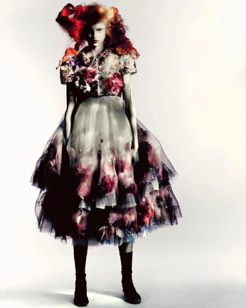 Fashion shot of a young female model in an ornate, layered tulle dress with floral details and striking headpiece, against a white backdrop.