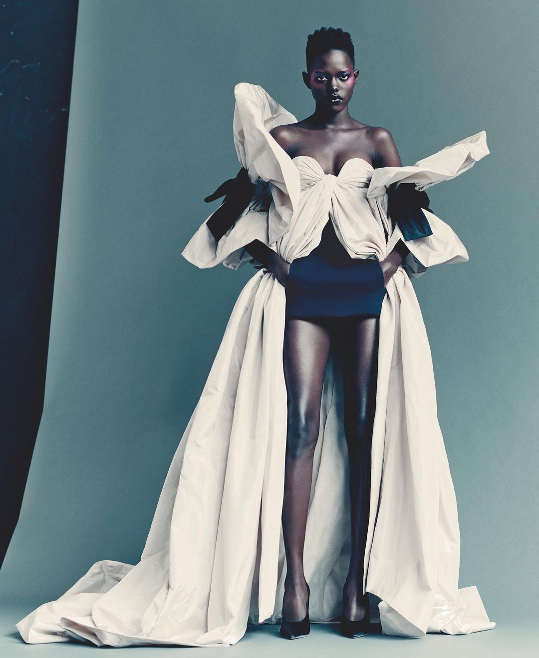 Glamorous fashion portrait of a Black woman in a couture gown with exaggerated ruffles, posing in a photo studio with a smooth grey backdrop.