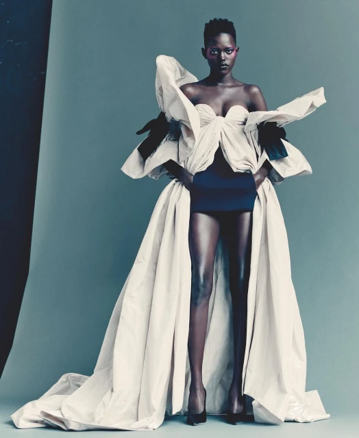 Glamorous fashion portrait of a Black woman in a couture gown with exaggerated ruffles, posing in a photo studio with a smooth grey backdrop.
