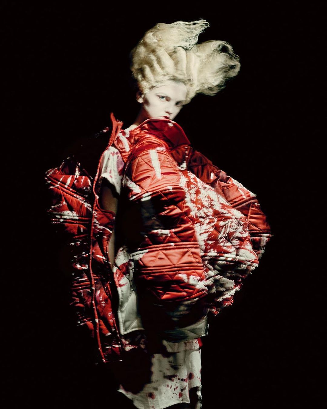 Fashion shot of a woman with an ornate hairstyle wearing a puffy red and white patterned coat in a dark studio setting.