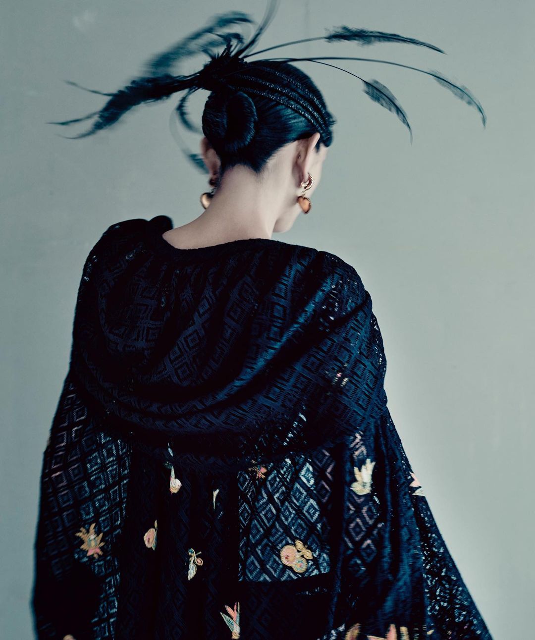 Back view of a person with braided updo adorned with feathers, wearing a black lace top with floral and geometric patterns.