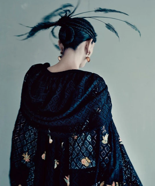 Back view of a person with braided updo adorned with feathers, wearing a black lace top with floral and geometric patterns.
