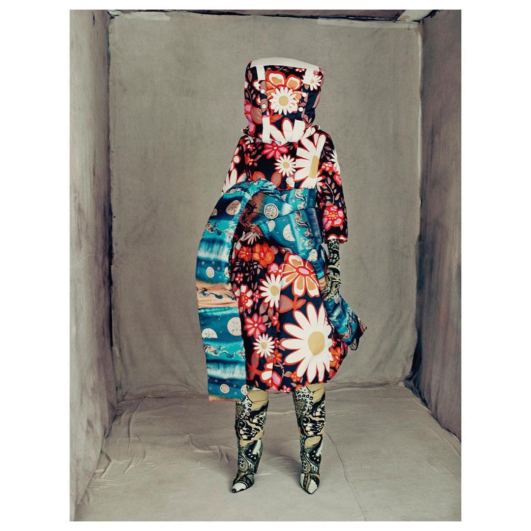 Person stands in a box-shaped room wearing a colorful floral coat with matching boots and gloves, creating a striking fashion statement.