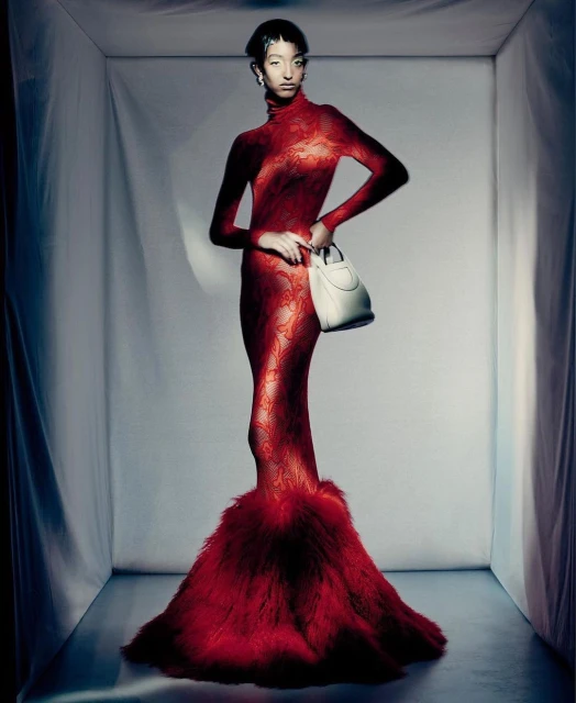 Full-length portrait of an adult woman in a red gown holding a white handbag, posing in a grey photo studio with white walls.