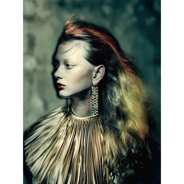 Indoor portrait of a woman with orange tinted hair wearing a gold pleated top, long earrings, and red lipstick posing in front of a textured wall.