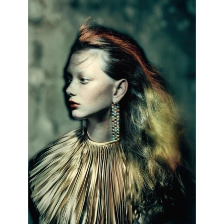 Indoor portrait of a woman with orange tinted hair wearing a gold pleated top, long earrings, and red lipstick posing in front of a textured wall.