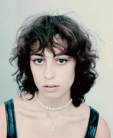 Portrait of a woman with dark curly hair wearing winged eyeliner, a pearl choker necklace, and a pendant necklace against a light background.