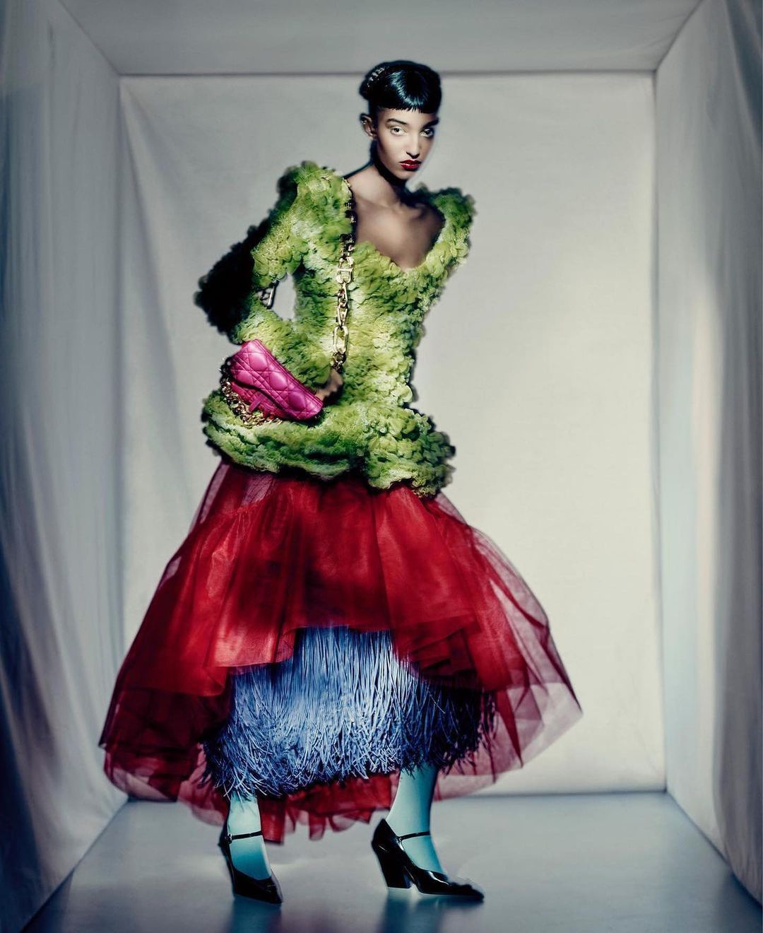 Elegant woman poses in a layered gown of tulle and faux-floral fabric with a bright pink purse against a stark white backdrop.