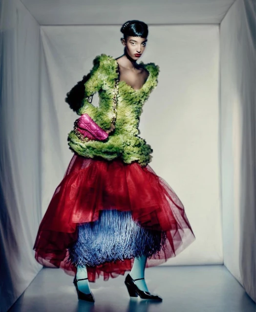 Elegant woman poses in a layered gown of tulle and faux-floral fabric with a bright pink purse against a stark white backdrop.
