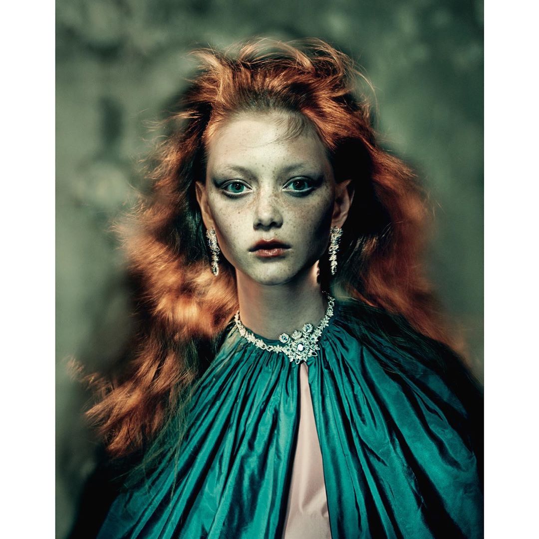 Close-up of a red-haired young woman with pale skin and freckles wearing jewelery and a teal cape in a fashion editorial photograph.
