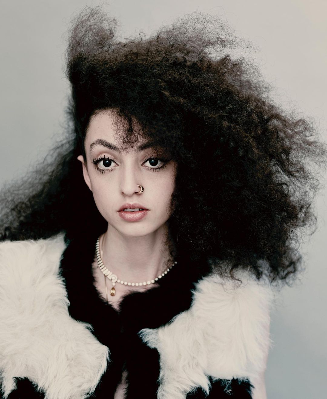 Close-up studio portrait of an adult female with curly afro hair wearing a fur vest and pearl necklace against a neutral grey background.