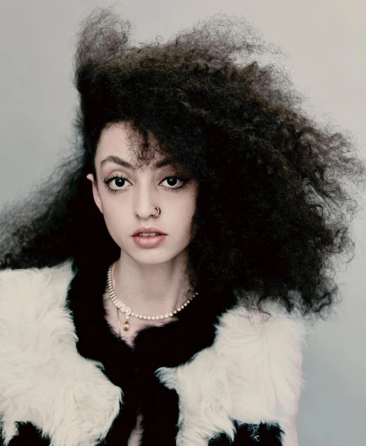 Close-up studio portrait of an adult female with curly afro hair wearing a fur vest and pearl necklace against a neutral grey background.