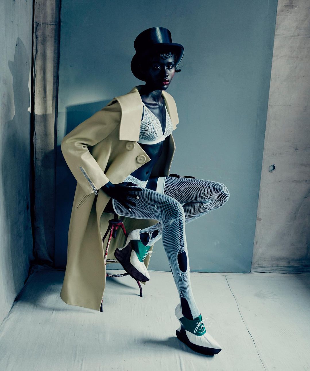 Dark-skinned fashion model poses on a stool wearing a top hat, bra top, ripped leggings, chunky shoes, and a light yellow jacket in a studio.