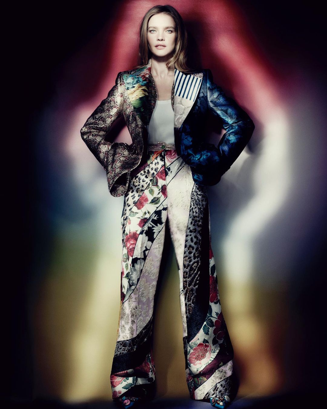 A woman stands in a brightly lit studio wearing a multicolor patchwork suit with floral and animal prints posing for a fashion editorial.