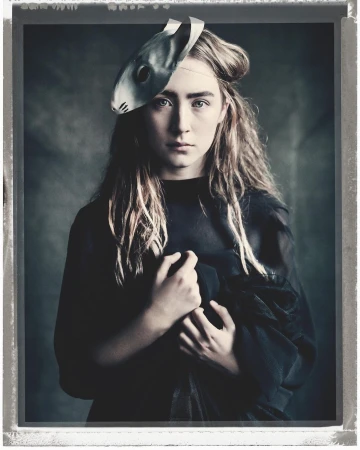 Dramatic portrait of a young woman wearing a mask with long hair, looking intently at the camera against a grey backdrop.
