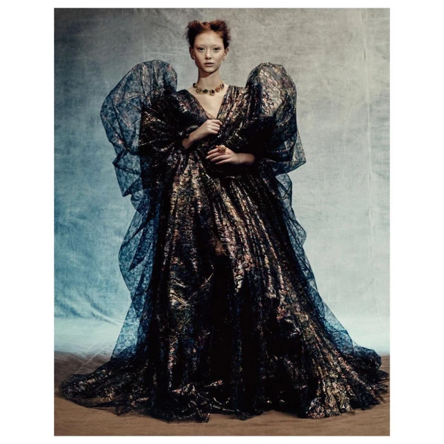 A fair-skinned woman in an elaborate black and gold gown with puffed sleeves stands against a mottled blue backdrop in a fashion shot.