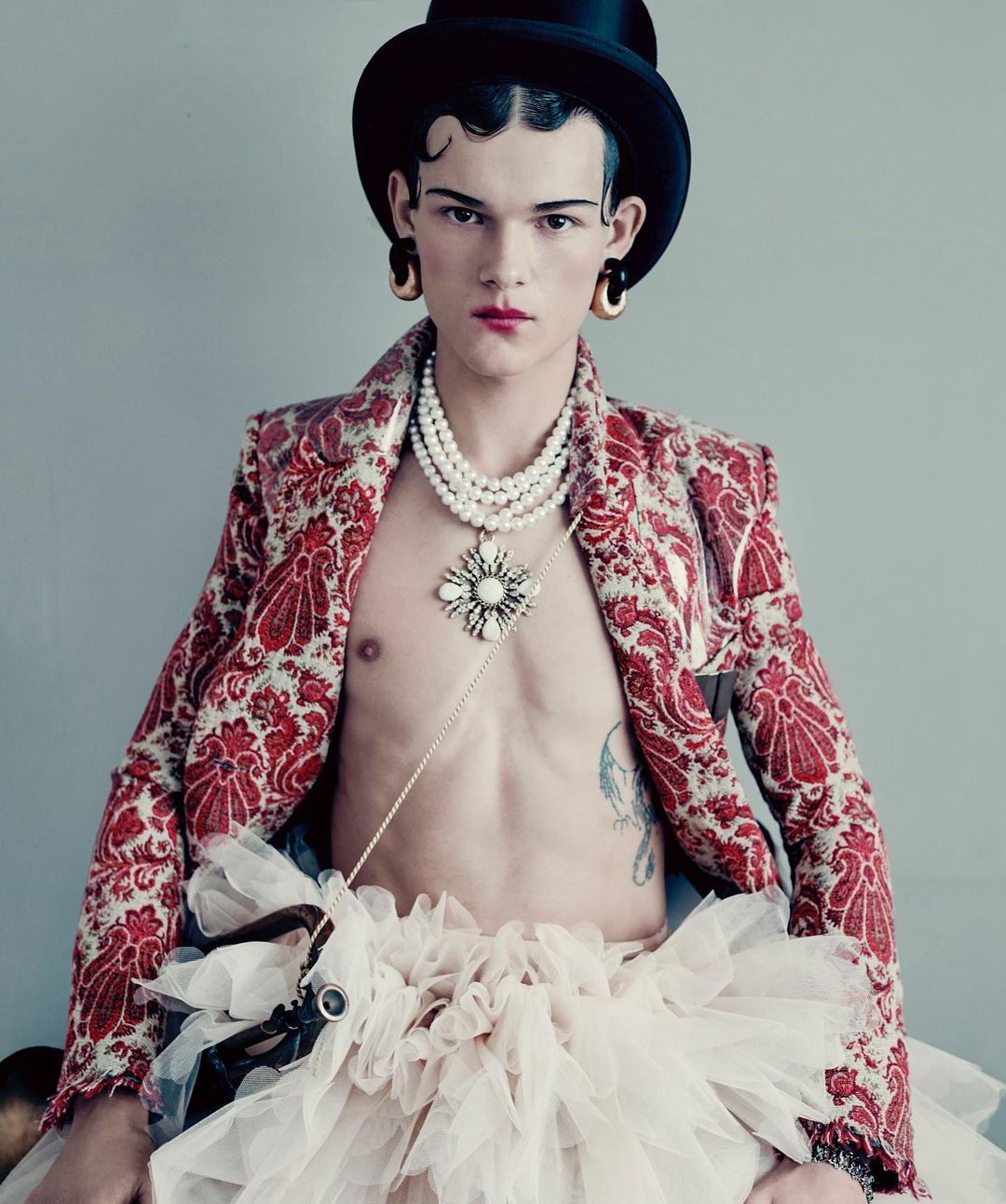 Fashionable young adult in red patterned jacket, top hat, pearl necklaces, and tutu, posing in a studio setting. Bold, artistic style.