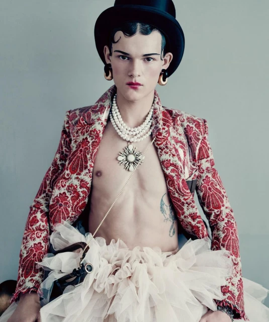 Fashionable young adult in red patterned jacket, top hat, pearl necklaces, and tutu, posing in a studio setting. Bold, artistic style.