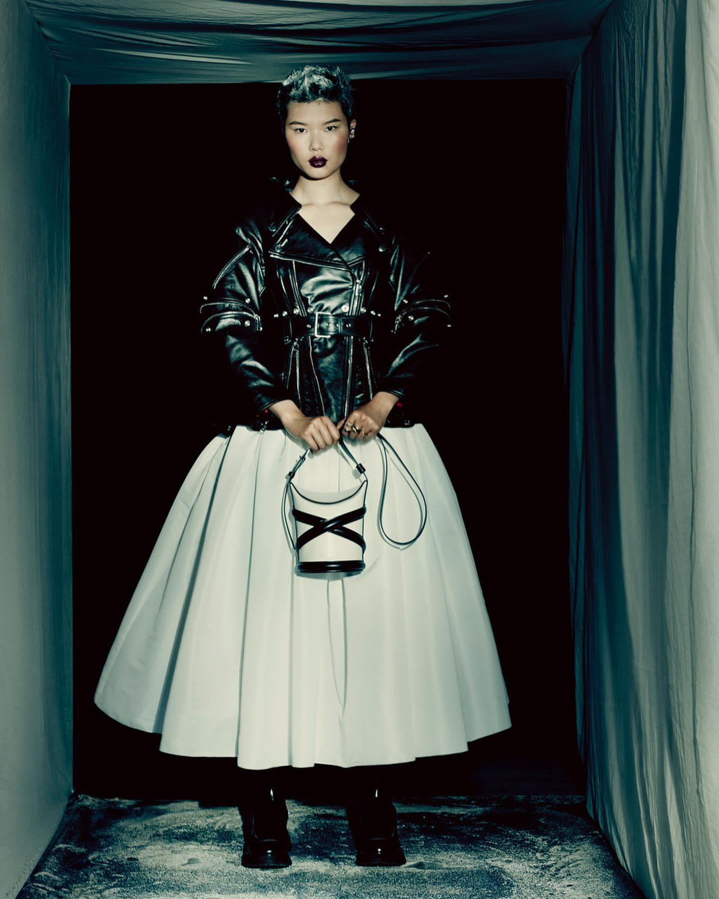 Full-length portrait of a fashionable Asian woman in a black leather jacket, white skirt, and boots, holding a bucket bag in a dark indoor space.