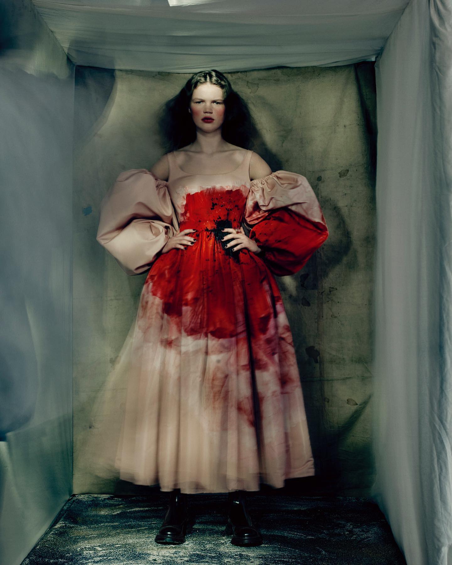 Fashion shot of a female model posing in a sculptural red-and-beige dress with puffed sleeves, set against a grey-blue studio backdrop.