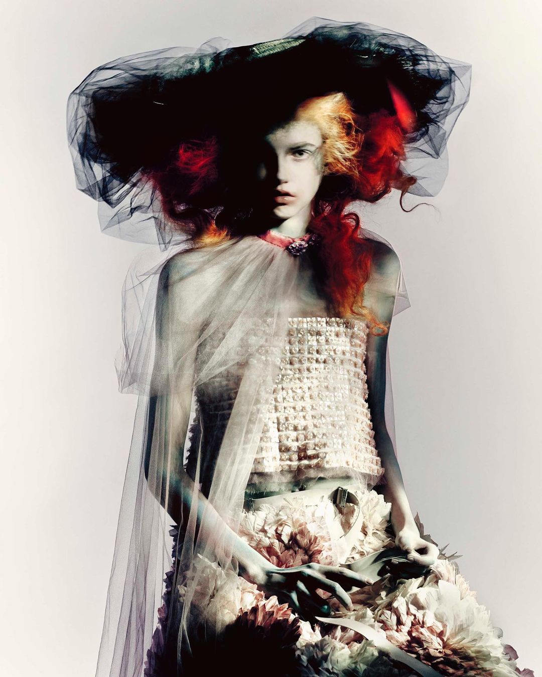 Fashionable young adult female model poses in a couture dress and oversized hat with red and blonde hair against a white background.