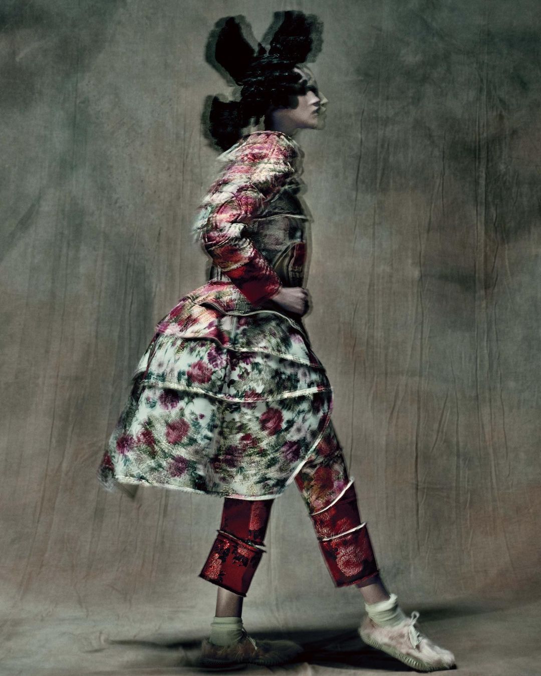 Model in profile wearing a floral print patterned coat and red patterned trousers against a textured mottled studio backdrop.