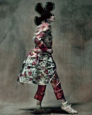 Model in profile wearing a floral print patterned coat and red patterned trousers against a textured mottled studio backdrop.