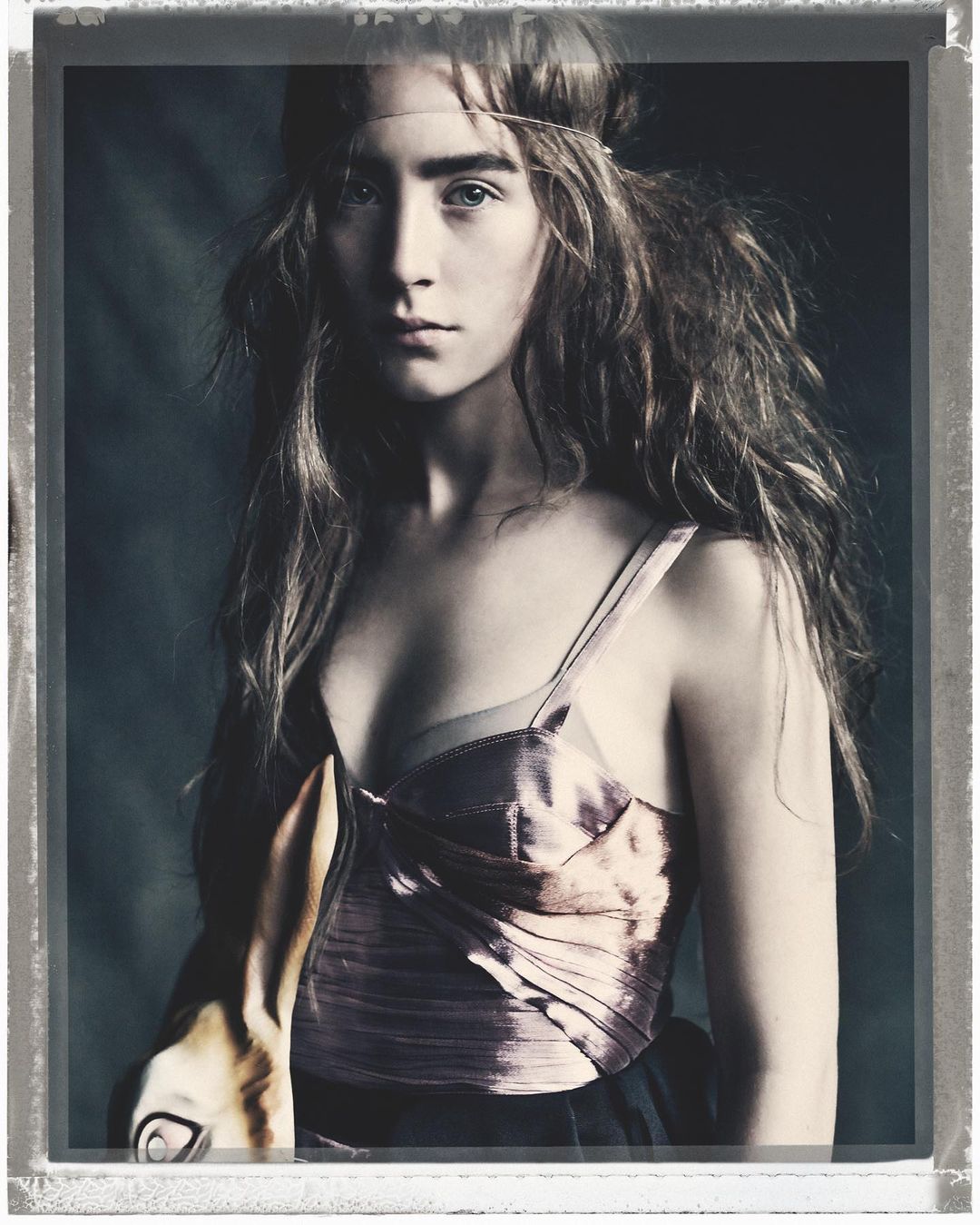 Portrait of a young woman wearing a pink corset top with long brown hair and a head band, inside a studio with a dark background.