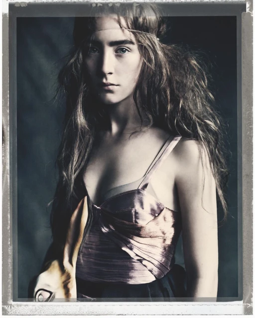 Portrait of a young woman wearing a pink corset top with long brown hair and a head band, inside a studio with a dark background.
