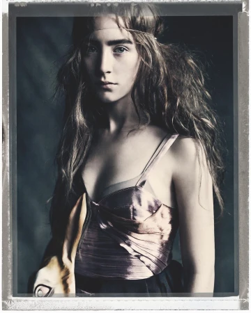 Portrait of a young woman wearing a pink corset top with long brown hair and a head band, inside a studio with a dark background.