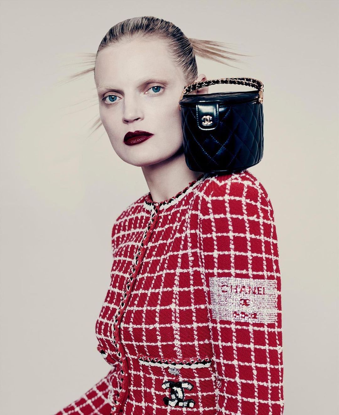 Woman in a red plaid jacket poses with a black quilted purse on her shoulder, pale skin, and dark red lipstick on a neutral background.